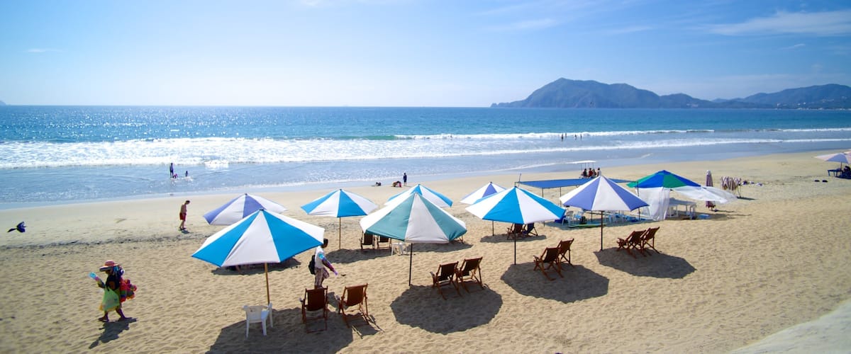 A beach with blue and white umbrellas, people, and a mountain in the background.