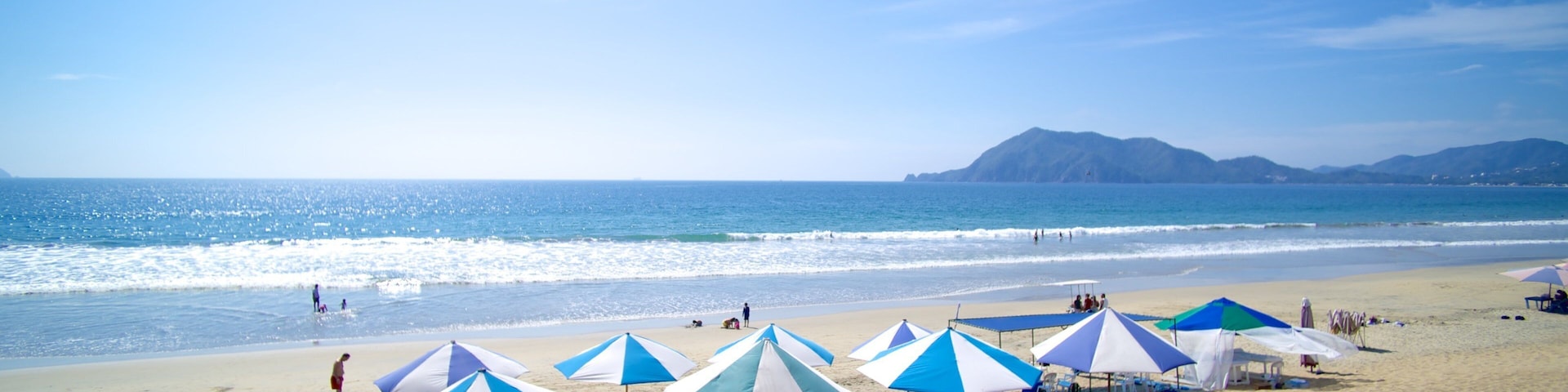 A beach with blue and white umbrellas, people, and a mountain in the background.