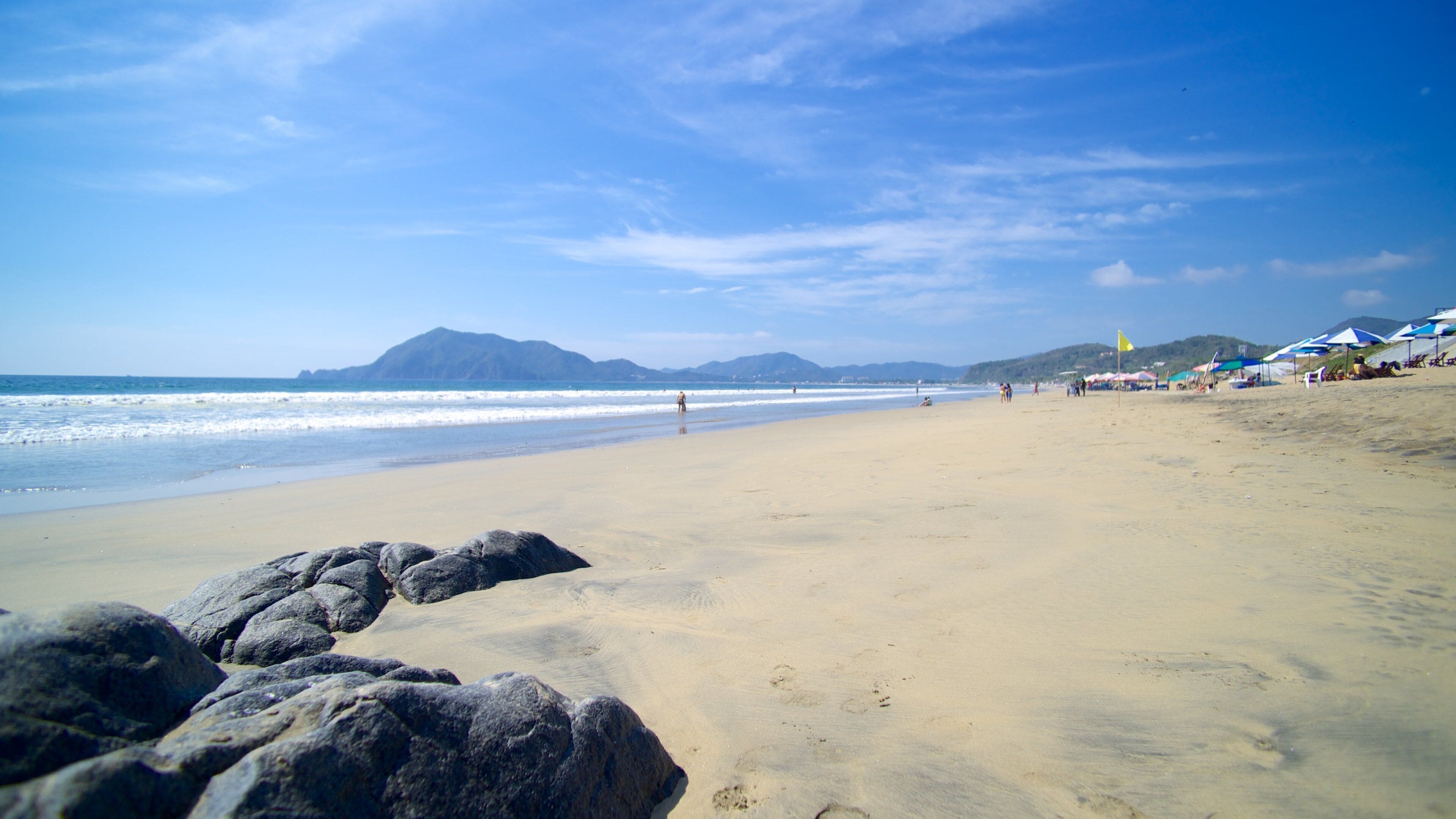 Una playa arenosa con rocas grandes al frente, olas rompiendo en la orilla y montañas a lo lejos.