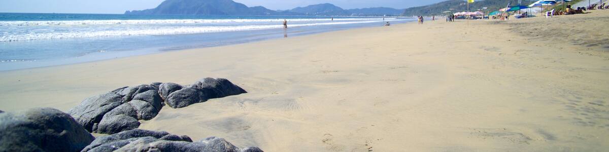 A sandy beach with large rocks in the foreground, waves crashing onto the shore, and mountains in the distance.