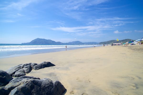A sandy beach with large rocks in the foreground, waves crashing onto the shore, and mountains in the distance.