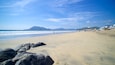 Ein sandiger Strand mit großen Felsen im Vordergrund, Wellen, die an die Küste branden, und Berge in der Ferne.