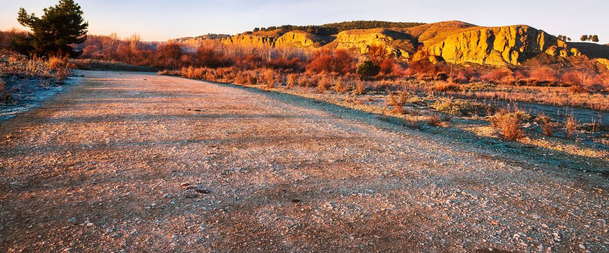 A gravel road leading through a dry, autumnal landscape with sparse vegetation and a prominent hill in the background.