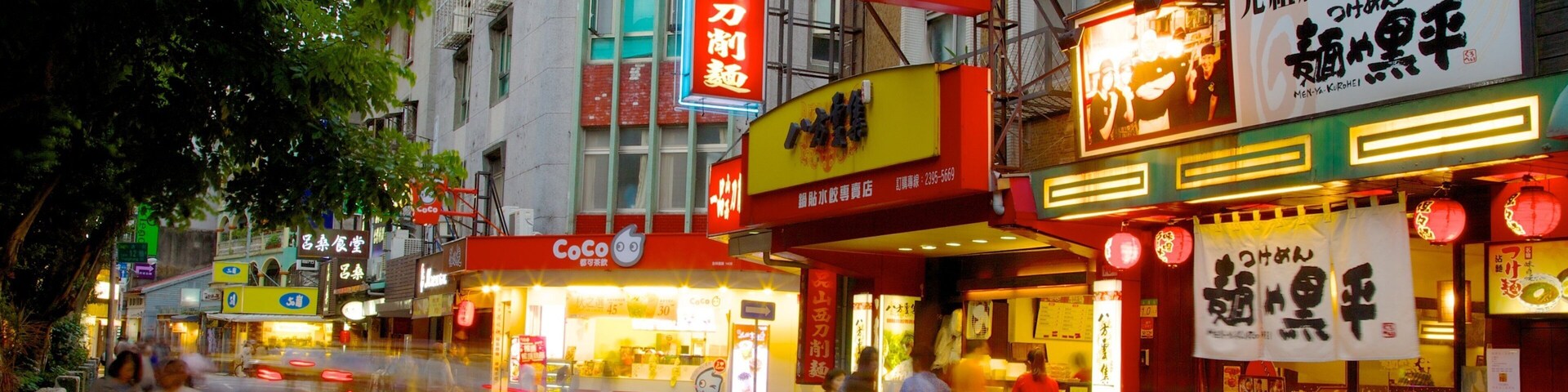 Rua movimentada com diversos estabelecimentos comerciais e placas, incluindo uma loja CoCo e um restaurante com letreiros chamativos.