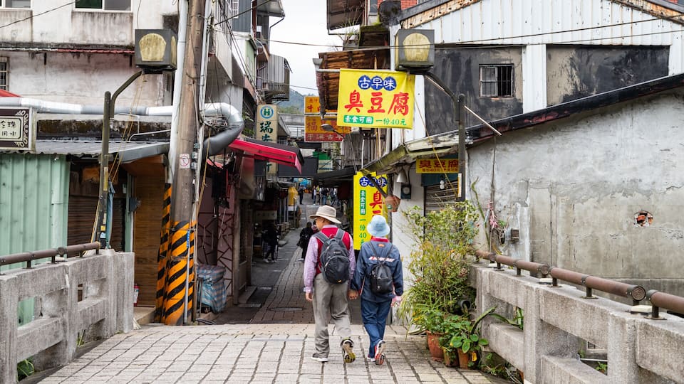 Uma viela estreita com duas pessoas caminhando, cercada por edifícios com placas em língua estrangeira.