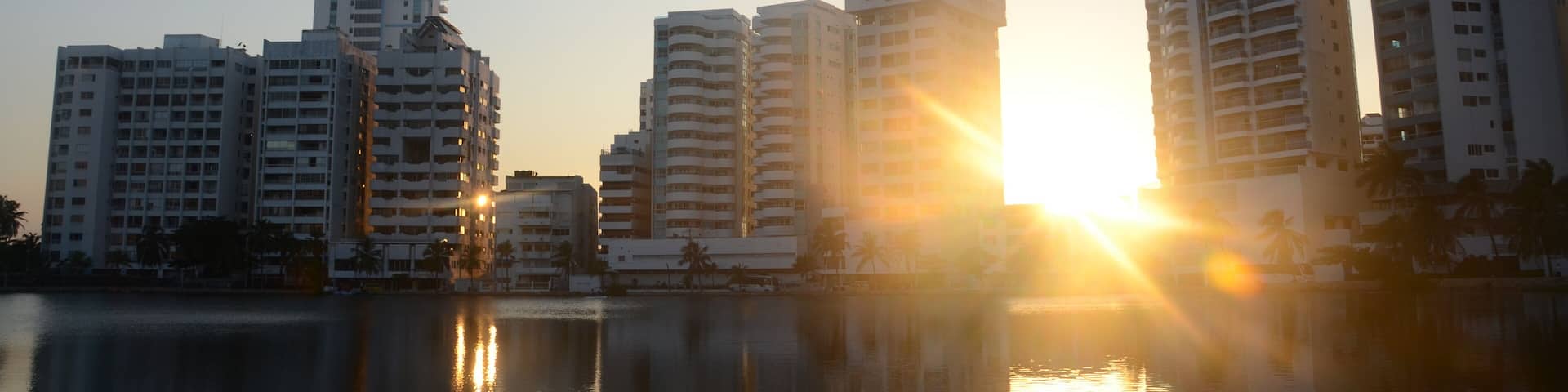 A city skyline with tall buildings reflecting on a calm body of water during sunset.
