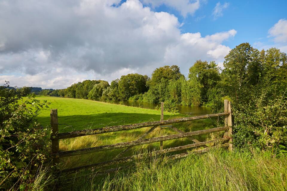 A rural landscape with a wooden fence, lush green fields, a river, and dense trees under a partly cloudy sky.