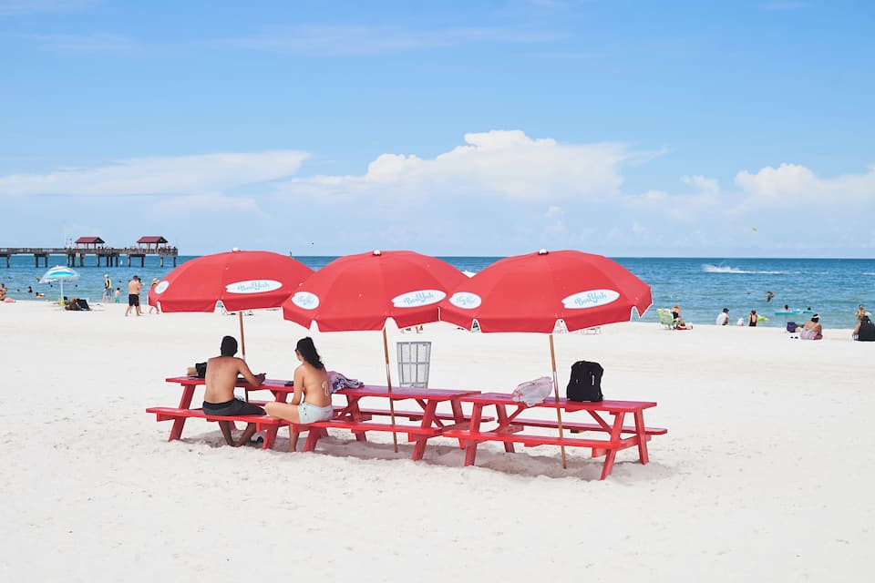 A beach with red umbrellas and picnic tables.