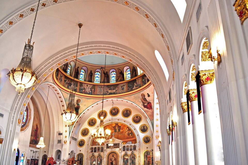 A church interior with ornate chandeliers, stained glass windows, and religious artwork on the walls.