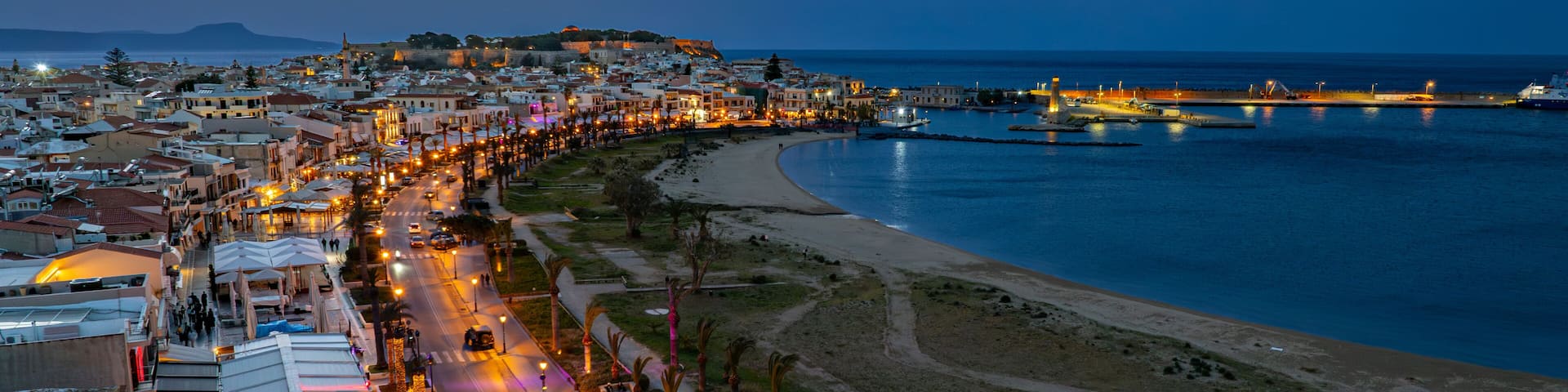 Aerial view of the Fortezza castle atop the hill, lights twinkling along the coastline, and the dark blue sea stretching out to the horizon, Rethimno, Rethymno, Greece.
