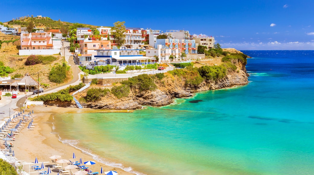 Sandy Varkotopos beach in sea bay of resort village Bali. Views of shore, washed by waves with sun loungers and parasols where sunbathing tourists. Greek houses stand on the rocky shore. Crete, Greece