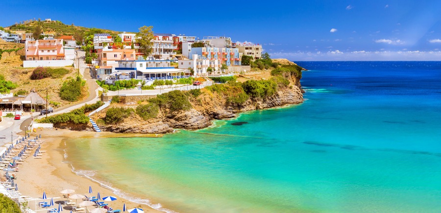 Sandy Varkotopos beach in sea bay of resort village Bali. Views of shore, washed by waves with sun loungers and parasols where sunbathing tourists. Greek houses stand on the rocky shore. Crete, Greece