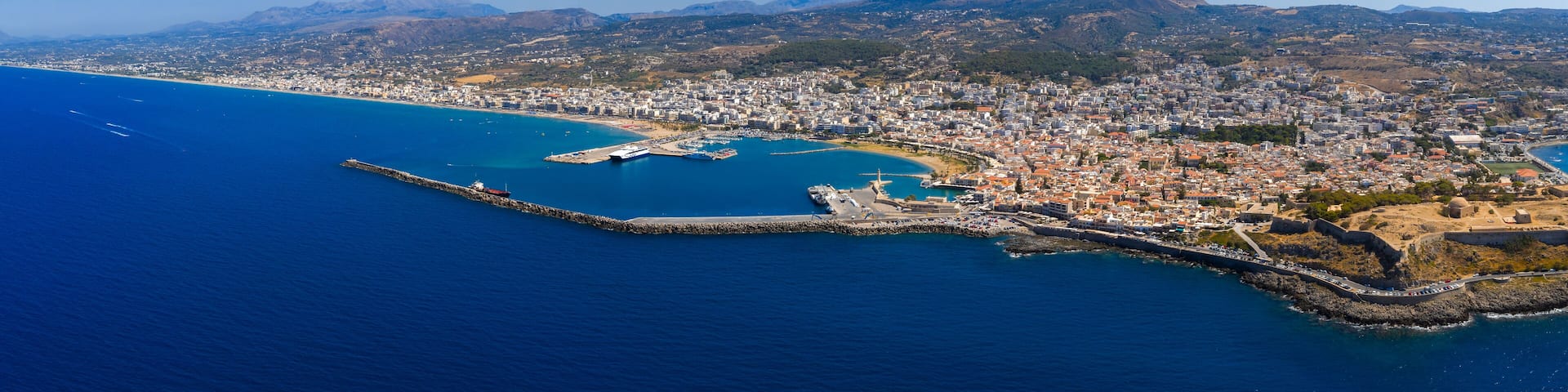 Aerial view of Rethymno town in Crete, Greece, featuring the Fortezza, terracotta roofed old town, Mediterranean harbor, and rugged mountains.
