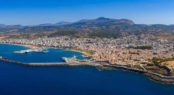 Vista aérea de una ciudad costera con una montaña imponente al fondo, un puerto con barcos y una zona fortificada con edificios históricos.