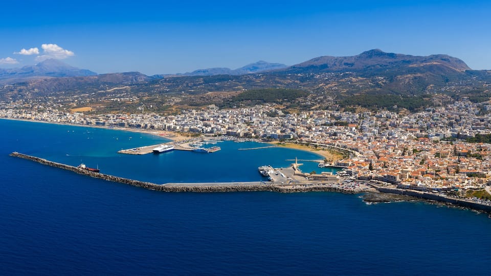 An aerial view of a coastal city with a prominent mountain in the background, a harbor with ships, and a fortified area with historical buildings.