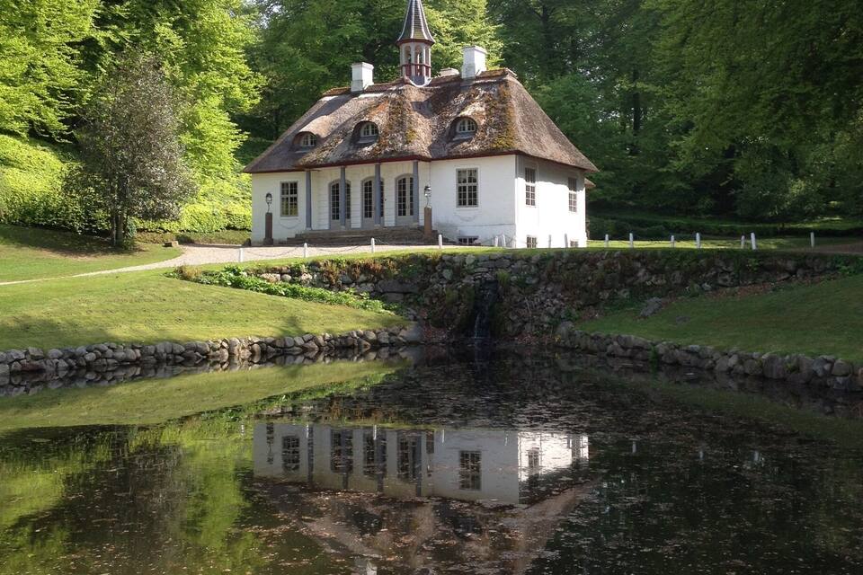 A white house with a thatched roof and a small tower, surrounded by lush greenery and a pond reflecting the structure.