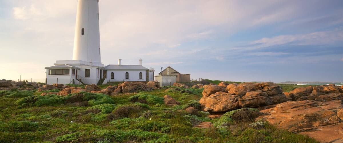 A white lighthouse with a small adjacent building, surrounded by rocky terrain and green vegetation.