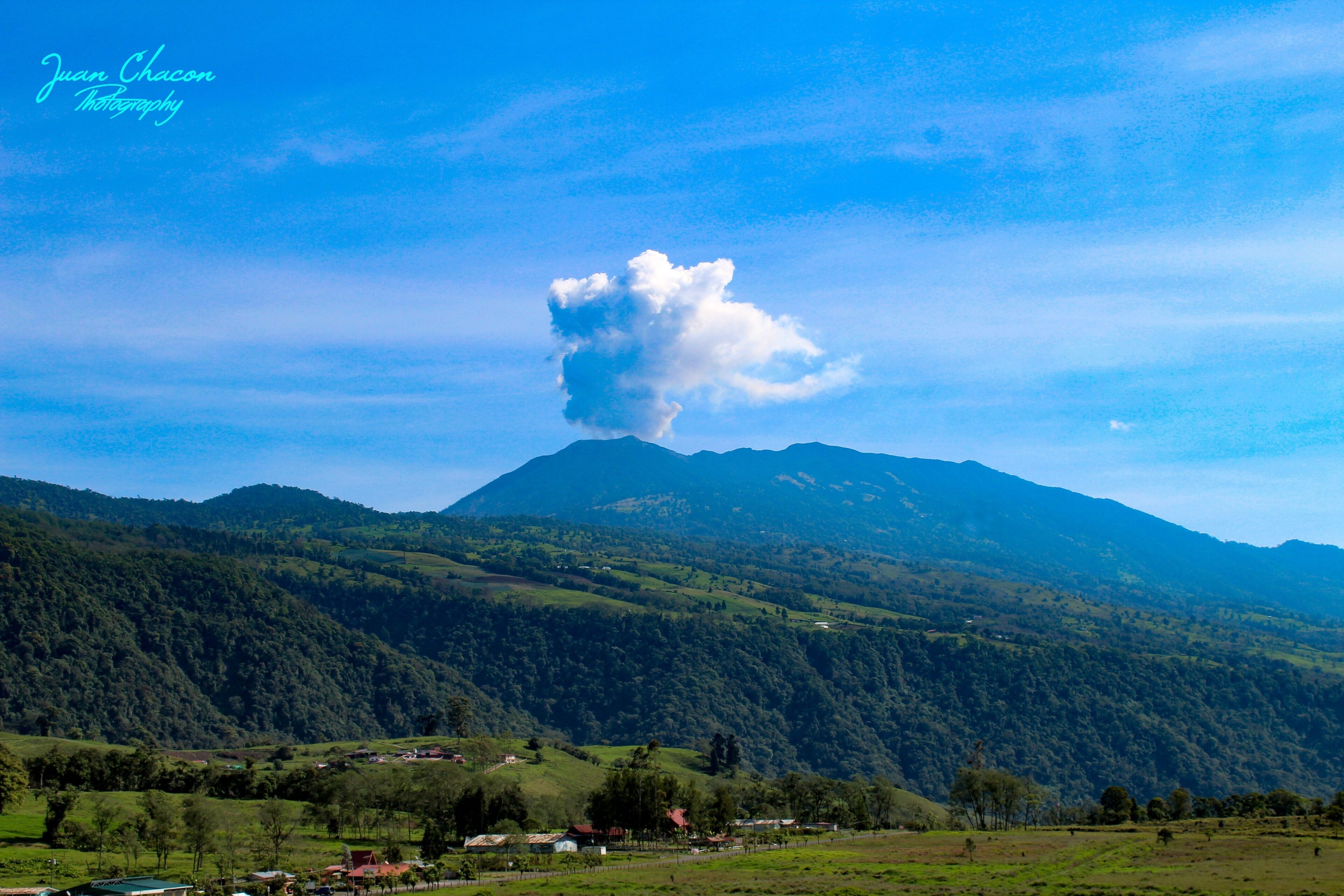 A mountain with a cloud in the sky, green hills, and a few houses.