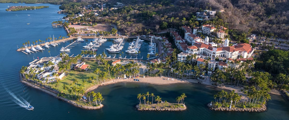 A marina surrounded by palm trees and houses on a coastal area with mountains in the background.
