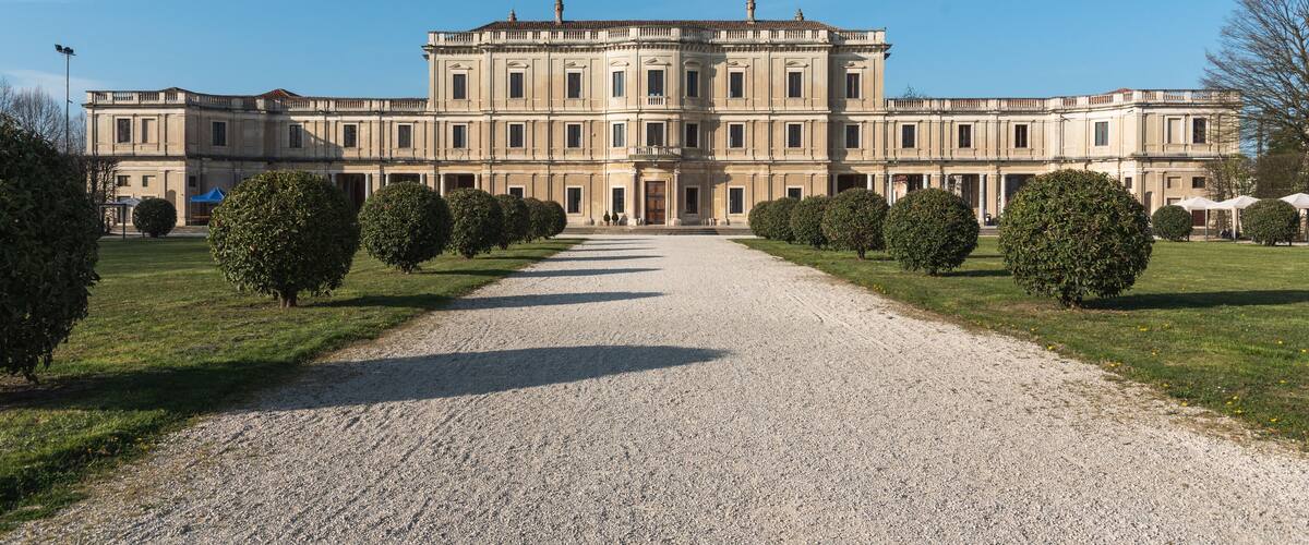 A large, classical building with a symmetrical facade, a gravel pathway leading to the entrance, and well-maintained hedges on either side.