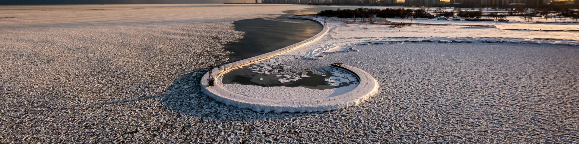 Beautiful Chicago skyline aerial drone view from above the frozen ice and snow covering Lake Michigan with a curved hooked pier near Montrose beach with colorful orange and pink sunset clouds above.