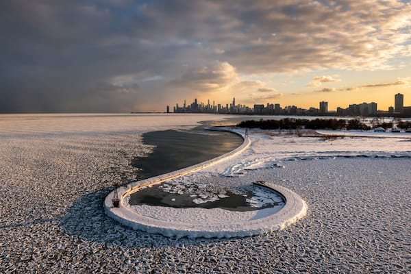 Beautiful Chicago skyline aerial drone view from above the frozen ice and snow covering Lake Michigan with a curved hooked pier near Montrose beach with colorful orange and pink sunset clouds above.