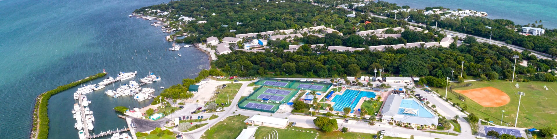 Vue aérienne d’une zone côtière comprenant un port de plaisance, des courts de tennis, un terrain de baseball et une végétation luxuriante environnante.