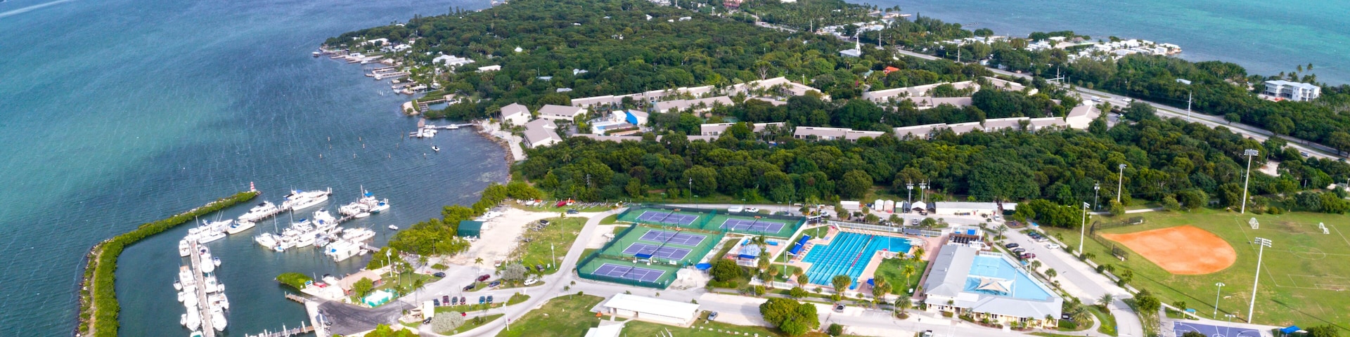 Vista aérea de una zona costera con un puerto deportivo, canchas de tenis, un campo de béisbol y vegetación circundante.