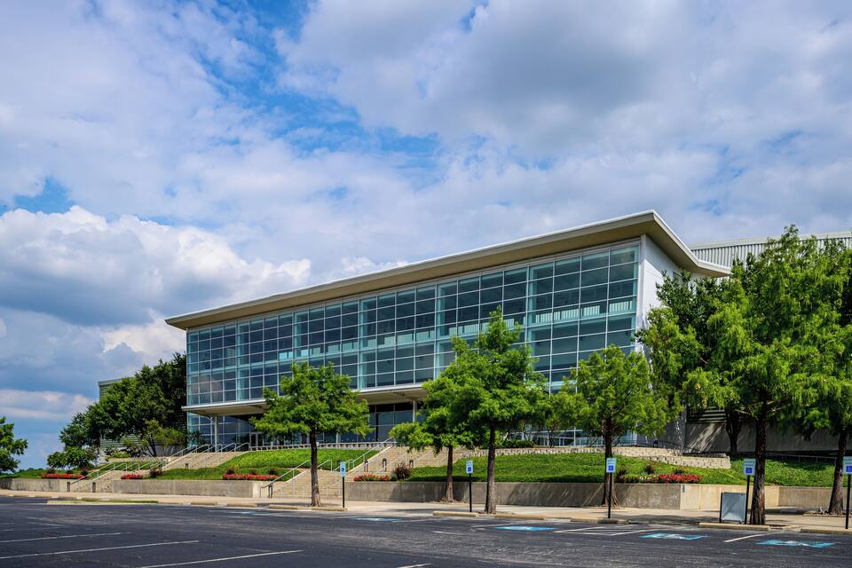 A modern building with large glass windows and a sloped roof, surrounded by trees and a parking lot.