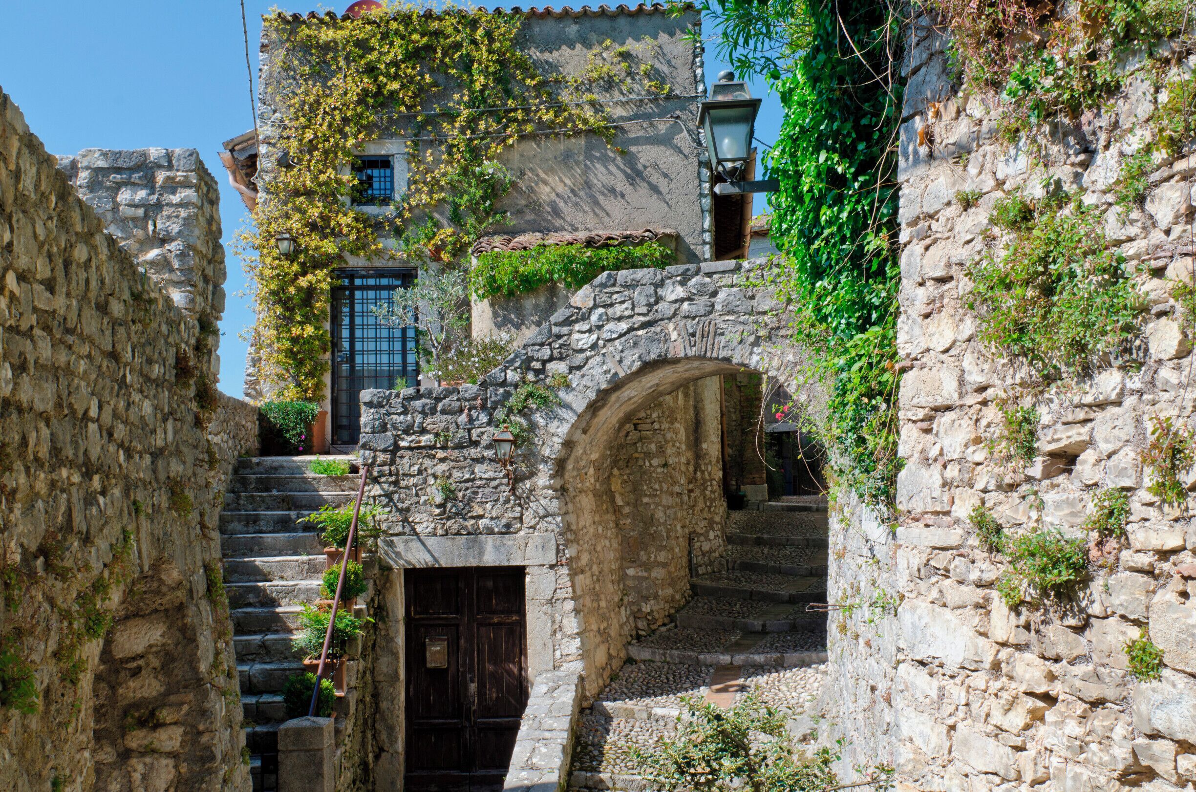 A stone staircase leading up to a building with a wooden door, surrounded by greenery and stone walls.