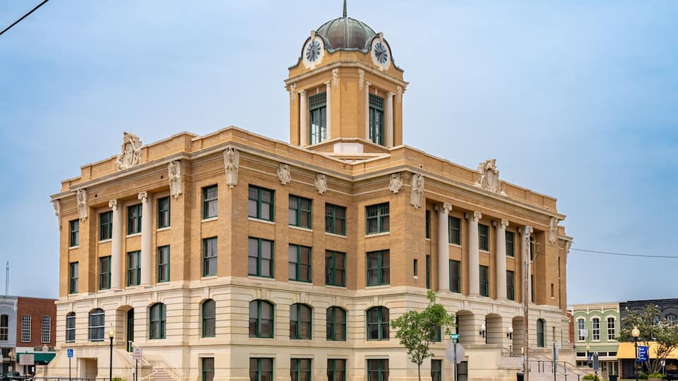 A large, historic building with a central dome and clock tower, surrounded by a well-maintained lawn and street.