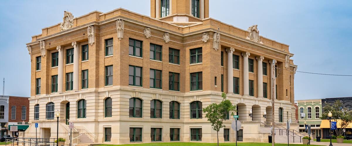 A large, historic building with a central dome and clock tower, surrounded by a well-maintained lawn and street.