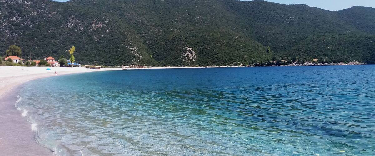 A clear beach with turquoise water, white sand, and green hills in the background.