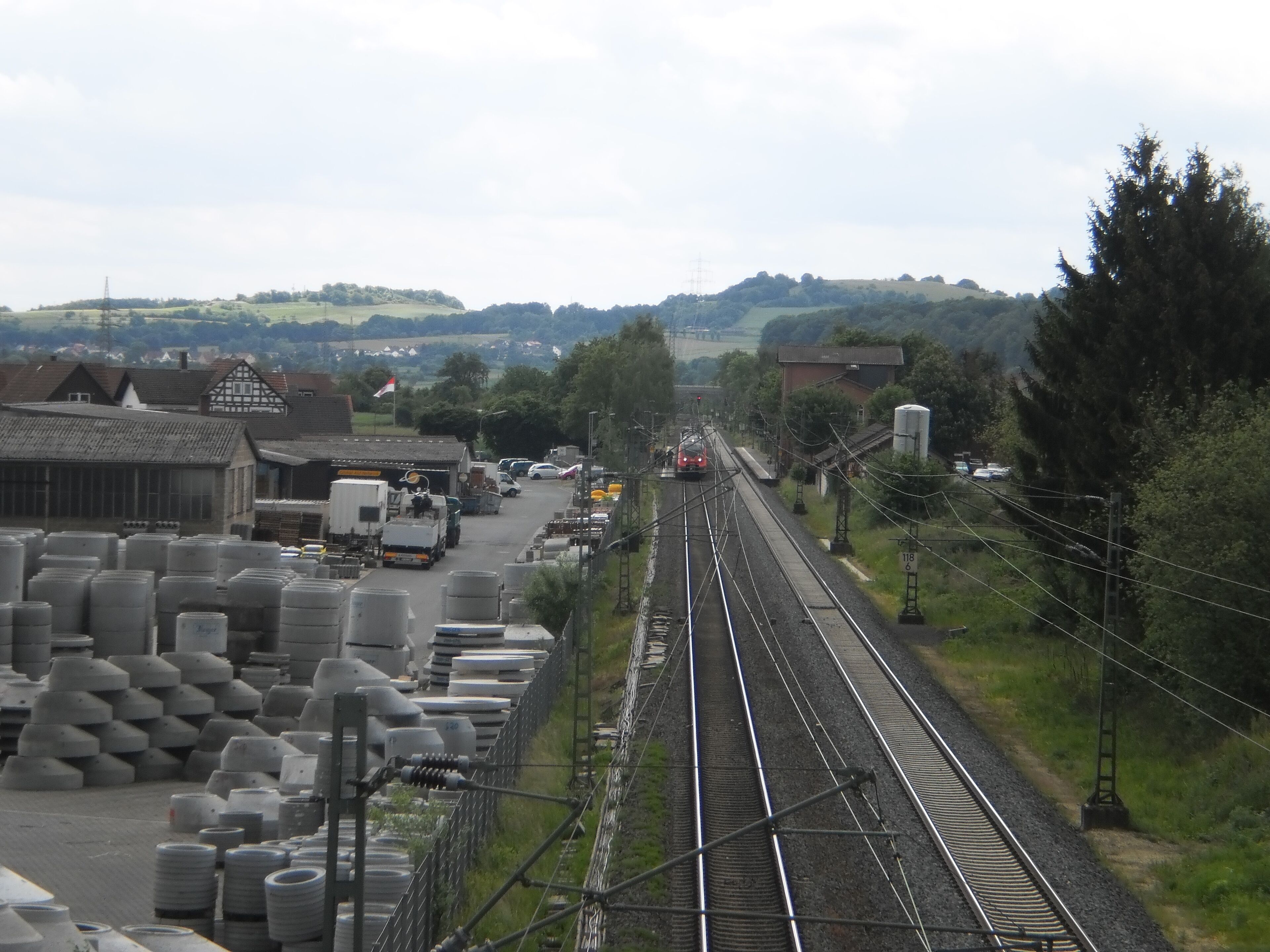 Blick von der Strassenbrücke der Ortsumfahrung von Fronhausen über die Main-Weser-Bahn auf den Bahnhof Fronhausen an der Lahn. Dort am Bahnsteig haltender Zug in Richtung Marburg und in den Landkreis Marburg-Biedenkopf nach Norden durch das Lahntal zwischen Gießen und Marburg. Links der Oberleitungen das Betonwerk Finger mit Röhren für Zisternen. In der Ferne ist von hier lahnabwärts der Altenberg oberhalb von Odenhausen zu sehen (bereits im Nachbarlandkreis Giessen).