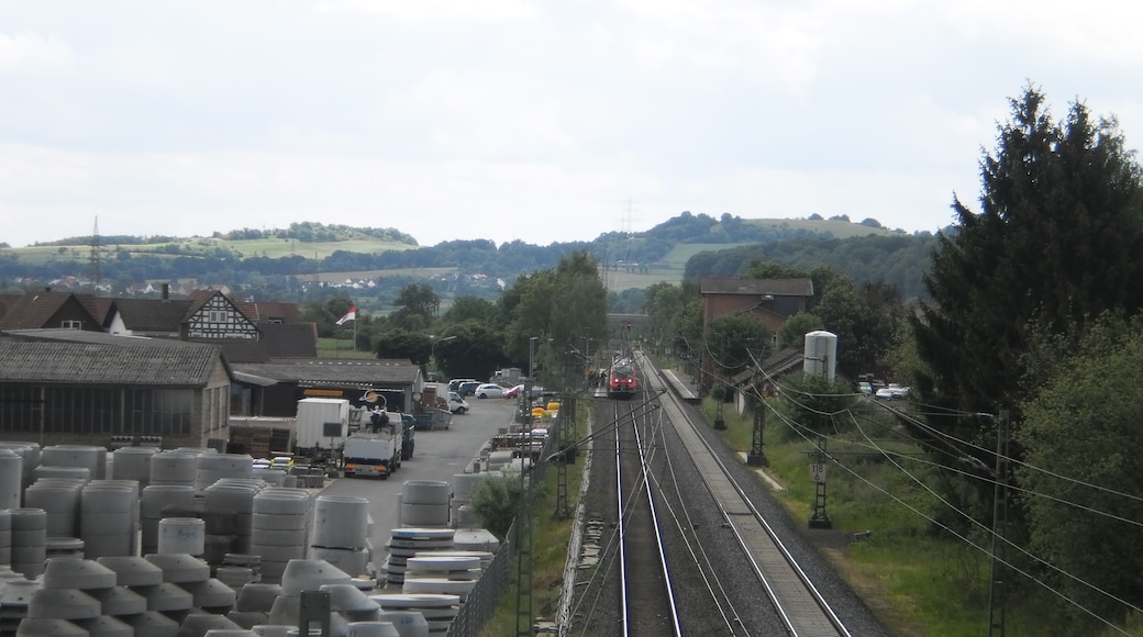 Blick von der Strassenbrücke der Ortsumfahrung von Fronhausen über die Main-Weser-Bahn auf den Bahnhof Fronhausen an der Lahn. Dort am Bahnsteig haltender Zug in Richtung Marburg und in den Landkreis Marburg-Biedenkopf nach Norden durch das Lahntal zwischen Gießen und Marburg. Links der Oberleitungen das Betonwerk Finger mit Röhren für Zisternen. In der Ferne ist von hier lahnabwärts der Altenberg oberhalb von Odenhausen zu sehen (bereits im Nachbarlandkreis Giessen).