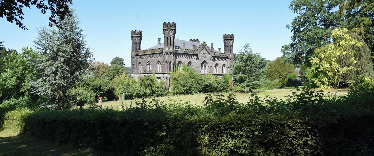 A large, historic building with multiple towers and a well-maintained garden in front.