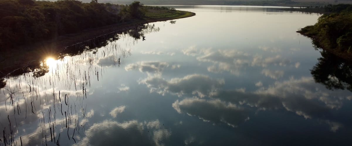 A calm river reflecting clouds, surrounded by dense vegetation and distant hills.