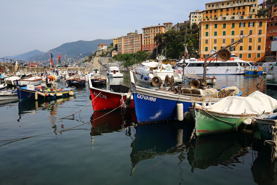 Un port esportiu amb diverses embarcacions amarrades, envoltat d'edificis de colors i un paisatge de muntanyes al fons.