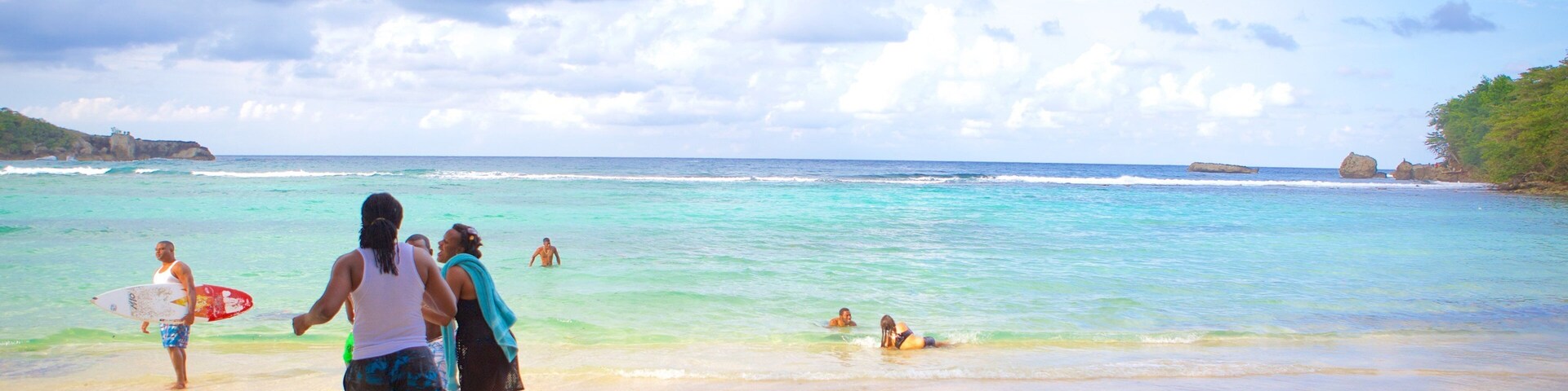 Una playa con agua turquesa cristalina, gente caminando y nadando, y afloramientos rocosos a lo lejos.