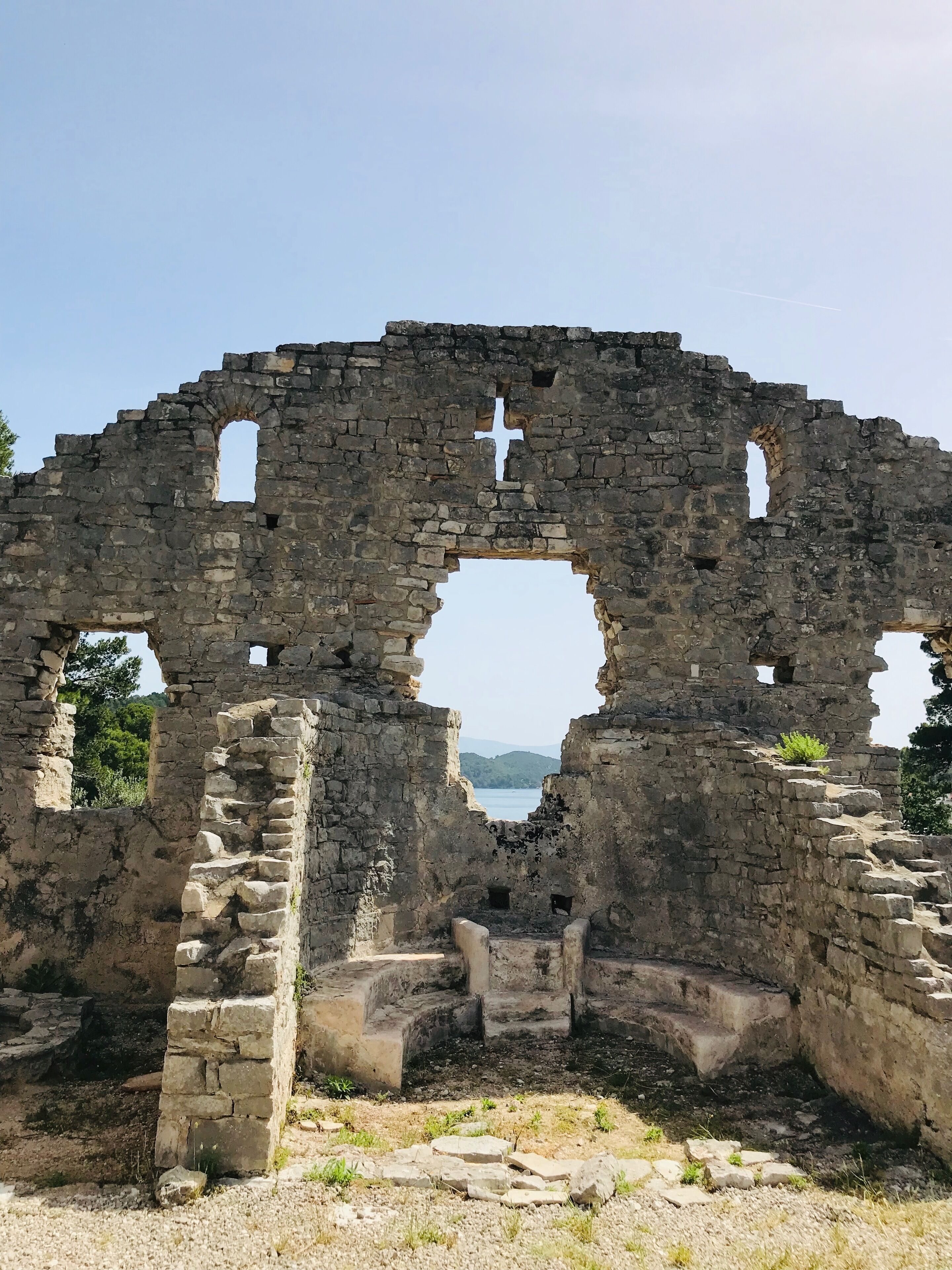 An ancient stone archway with a cross at the top, surrounded by ruins and a view of the sea.