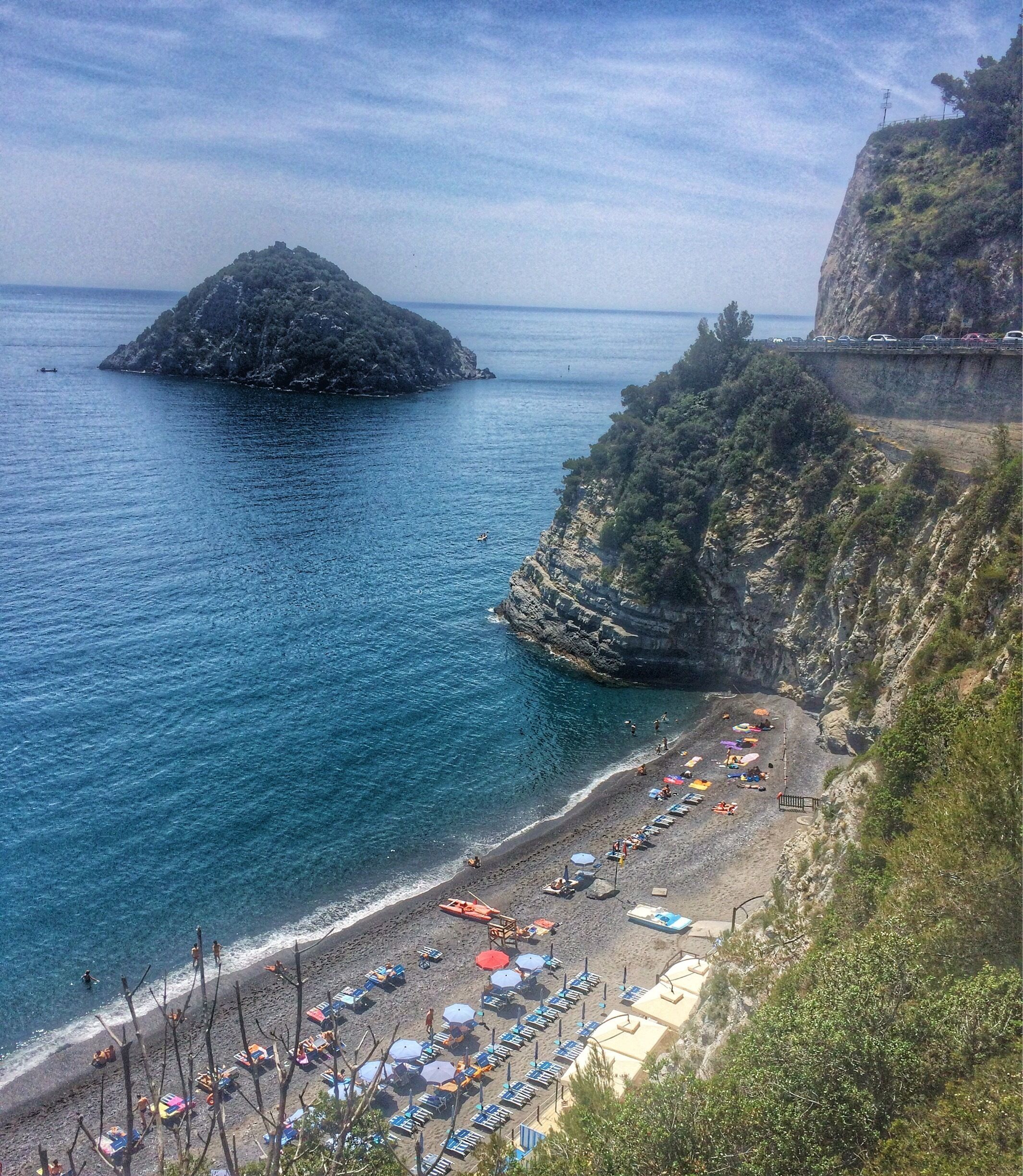 A coastal beach with umbrellas and sun loungers, rocky cliffs, and a small island in the water.