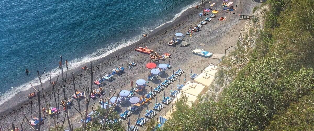 A coastal beach with umbrellas and sun loungers, rocky cliffs, and a small island in the water.