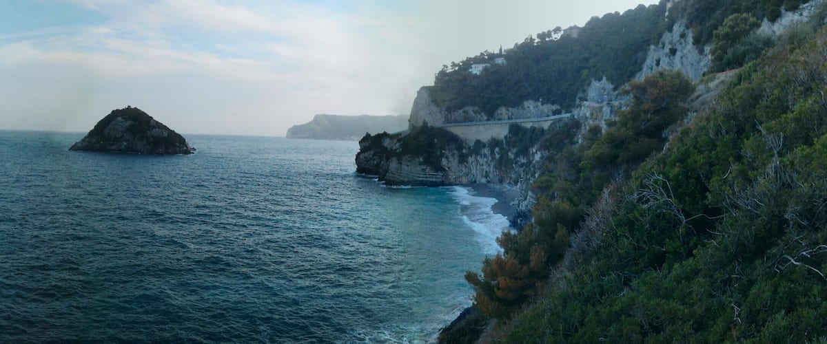 Paesaggio costiero con un promontorio roccioso, una piccola isola e un ponte che collega la terraferma a una scogliera.