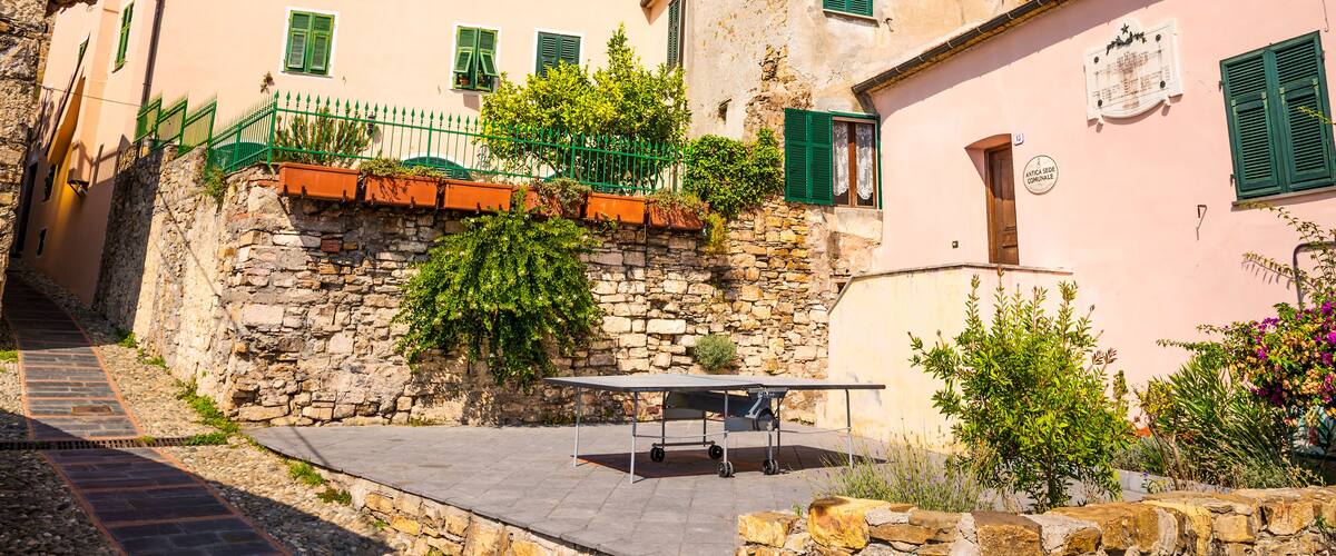 A narrow street with stone walls, a table, and buildings with green shutters.