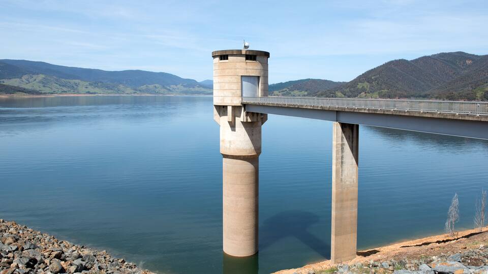 A bridge over a calm lake with a tower-like structure on one end and a rocky shoreline in the foreground.