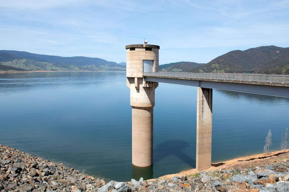 A bridge over a calm lake with a tower-like structure on one end and a rocky shoreline in the foreground.