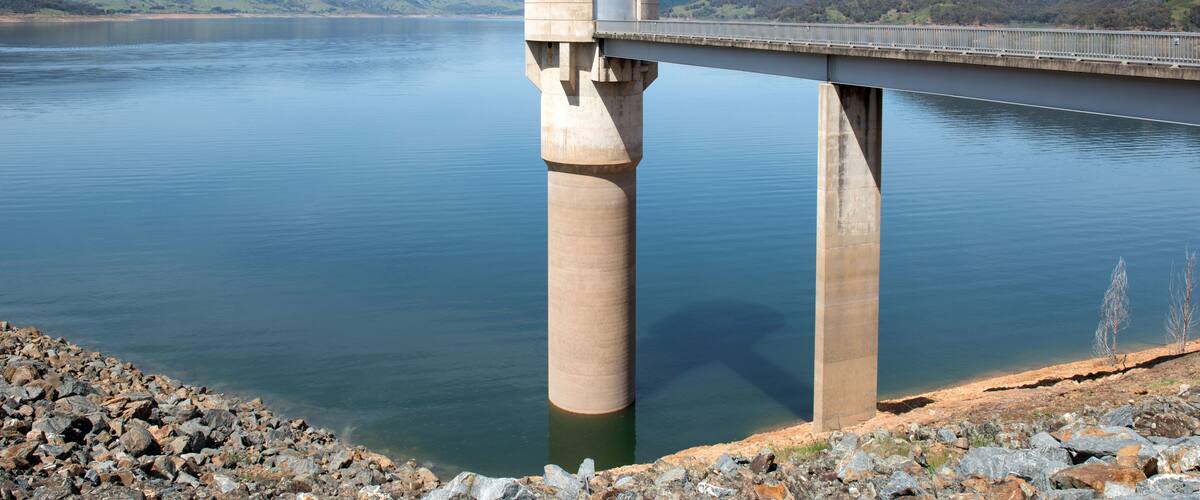 A bridge over a calm lake with a tower-like structure on one end and a rocky shoreline in the foreground.