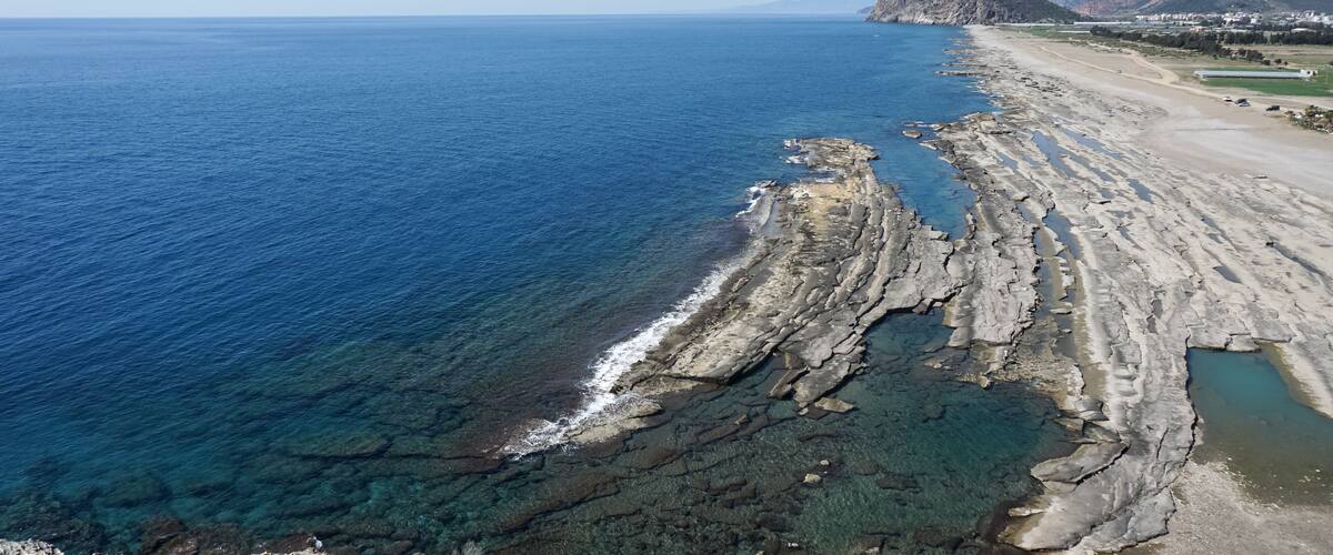 A coastal landscape with a rocky shoreline, clear blue sea, and distant mountains.