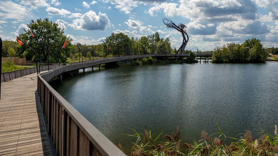 A wooden boardwalk over a calm lake with a metal sculpture and trees in the background.