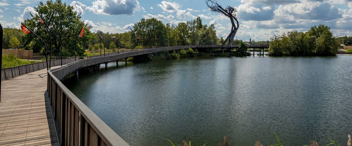A wooden boardwalk over a calm lake with a metal sculpture and trees in the background.